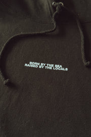 Black hoodie with text 'Born by the Sea, Raised by the Locals' on a dark background