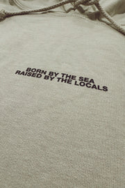 Text 'Born by the Sea, Raised by the Locals' on a beige hoodie.